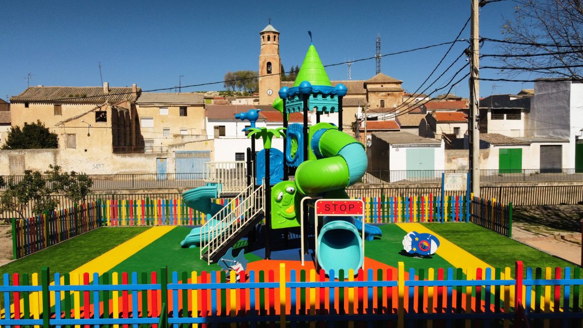 La imagen muestra un parque infantil vallado con una estructura de juegos colorida en el centro. La estructura tiene toboganes de diferentes formas, escaleras y elementos decorativos como una torre con techo en forma de cono verde, palmeras de plástico y un panel con una cara sonriente. La valla que rodea el parque es de colores brillantes (rojo, azul, amarillo y verde).