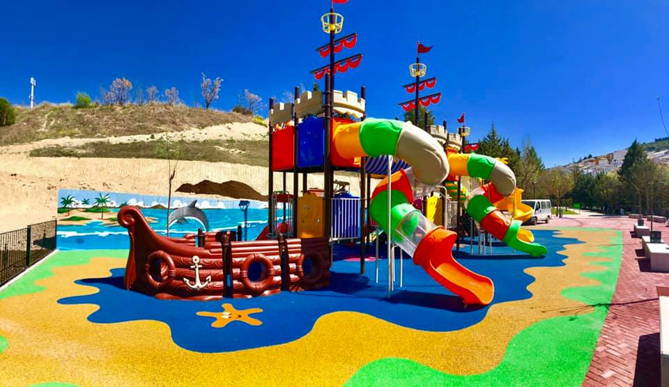 El parque infantil cuenta con un diseño temático inspirado en un barco pirata, ofreciendo una experiencia lúdica única para los niños. Dispone de una gran estructura con toboganes coloridos en forma de tubos, plataformas elevadas, torres con banderas y detalles que evocan un castillo. La zona de juego está decorada con un suelo de goma que simula el mar y la arena, proporcionando seguridad y un ambiente inmersivo. Además, un mural pintado en el fondo refuerza la temática marina con palmeras y paisajes tropicales. Es un espacio ideal para la diversión en un entorno seguro y atractivo.
