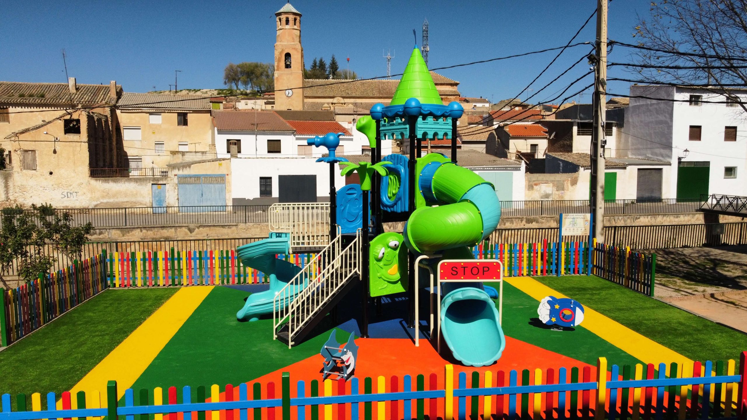 Parque infantil vallado con suelo de colores vibrantes y un gran conjunto de juegos con temática de castillo. La estructura principal es azul y verde, con toboganes, escaleras y plataformas de juego. Rodeado por una valla de madera pintada en colores llamativos, el parque está ubicado en una zona residencial con casas blancas y verdes, árboles y un puente en el fondo.