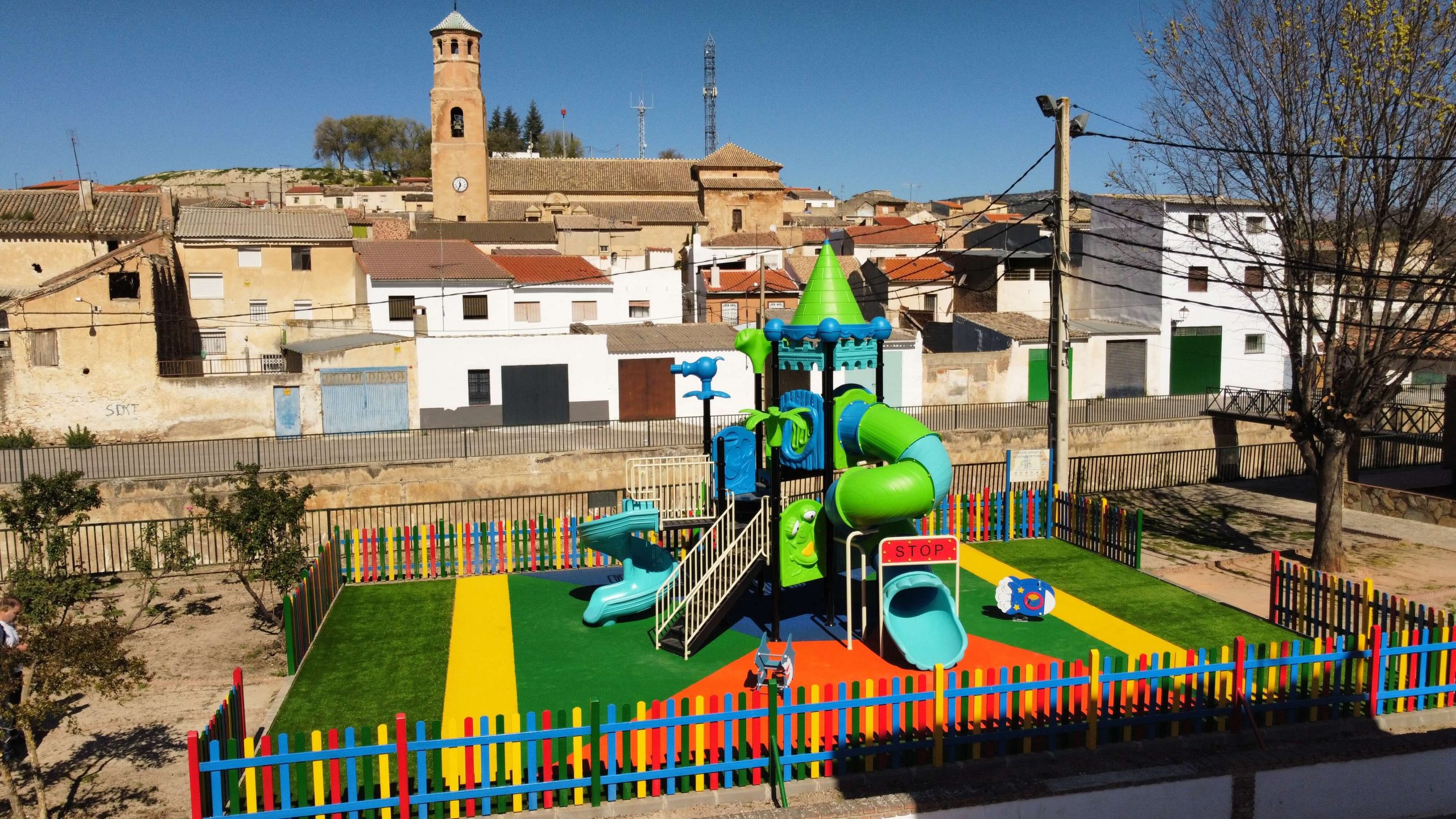 Parque infantil vallado con suelo de colores vibrantes y un gran conjunto de juegos con temática de castillo. La estructura principal es azul y verde, con toboganes, escaleras y plataformas de juego. Rodeado por una valla de madera pintada en colores llamativos, el parque está ubicado en una zona residencial con casas blancas y verdes, árboles y un puente en el fondo.
