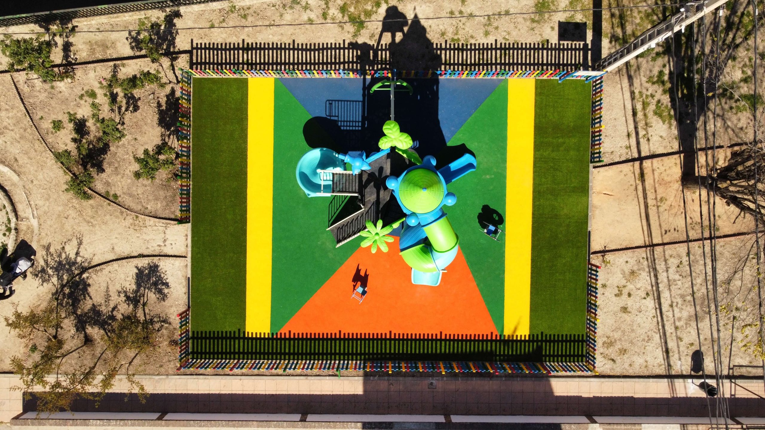 dji_fly_20240412_113900_500_1712922004839_photo_optimized Parque infantil vallado con suelo de colores vibrantes y un gran conjunto de juegos con temática de castillo. La estructura principal es azul y verde, con toboganes, escaleras y plataformas de juego. Rodeado por una valla de madera pintada en colores llamativos, el parque está ubicado en una zona residencial con casas blancas y verdes, árboles y un puente en el fondo.