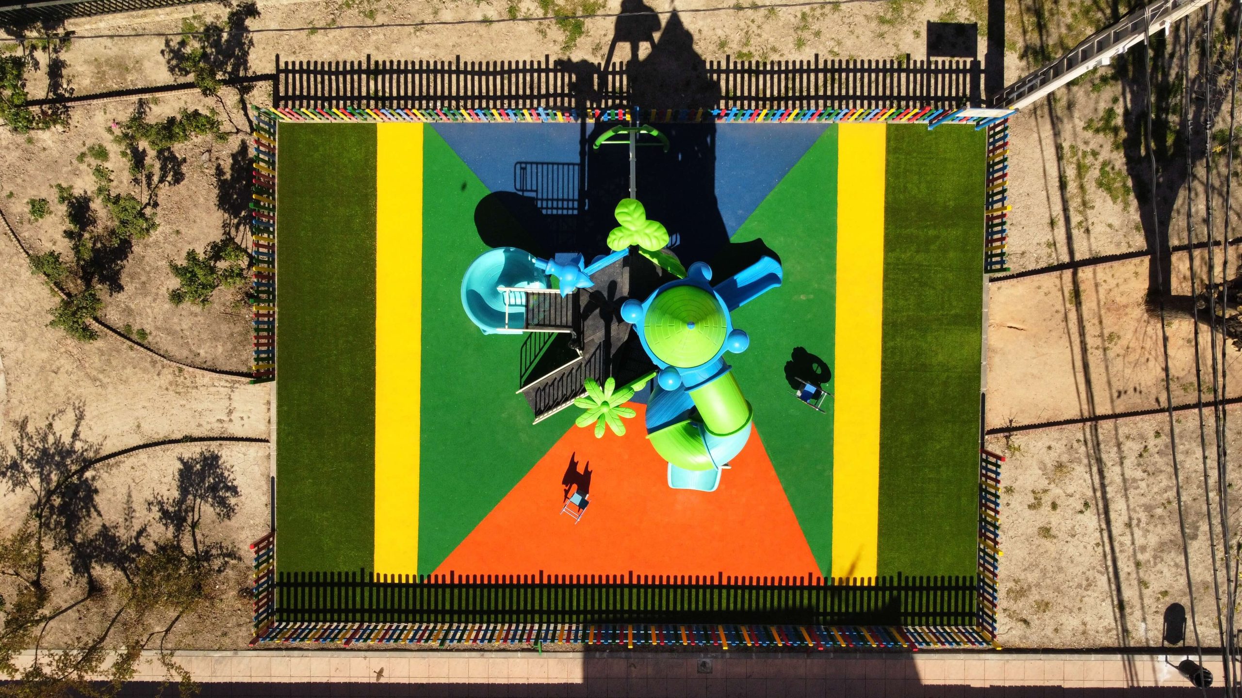 Parque infantil vallado con suelo de colores vibrantes y un gran conjunto de juegos con temática de castillo. La estructura principal es azul y verde, con toboganes, escaleras y plataformas de juego. Rodeado por una valla de madera pintada en colores llamativos, el parque está ubicado en una zona residencial con casas blancas y verdes, árboles y un puente en el fondo.