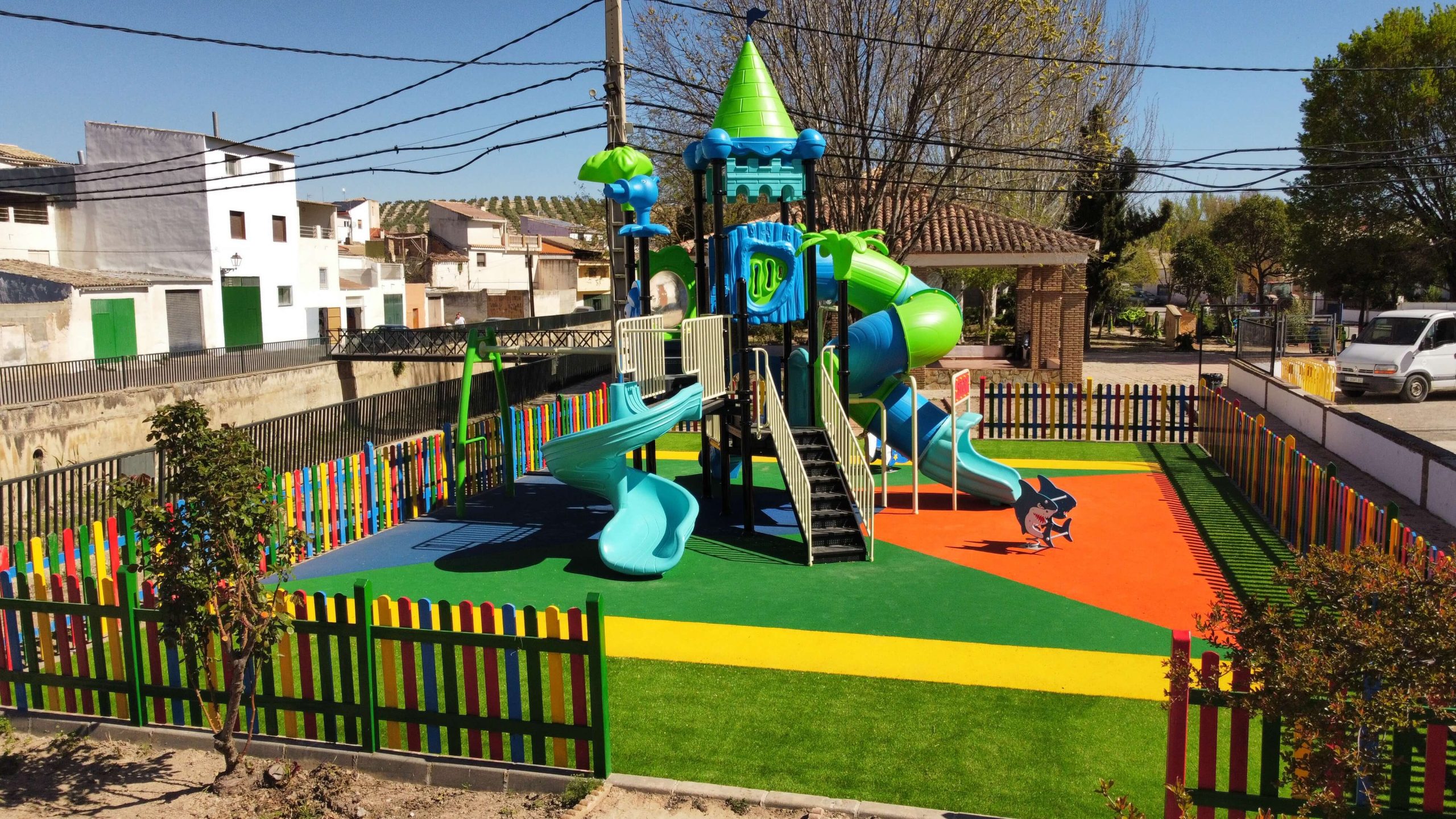 Parque infantil vallado con suelo de colores vibrantes y un gran conjunto de juegos con temática de castillo. La estructura principal es azul y verde, con toboganes, escaleras y plataformas de juego. Rodeado por una valla de madera pintada en colores llamativos, el parque está ubicado en una zona residencial con casas blancas y verdes, árboles y un puente en el fondo.