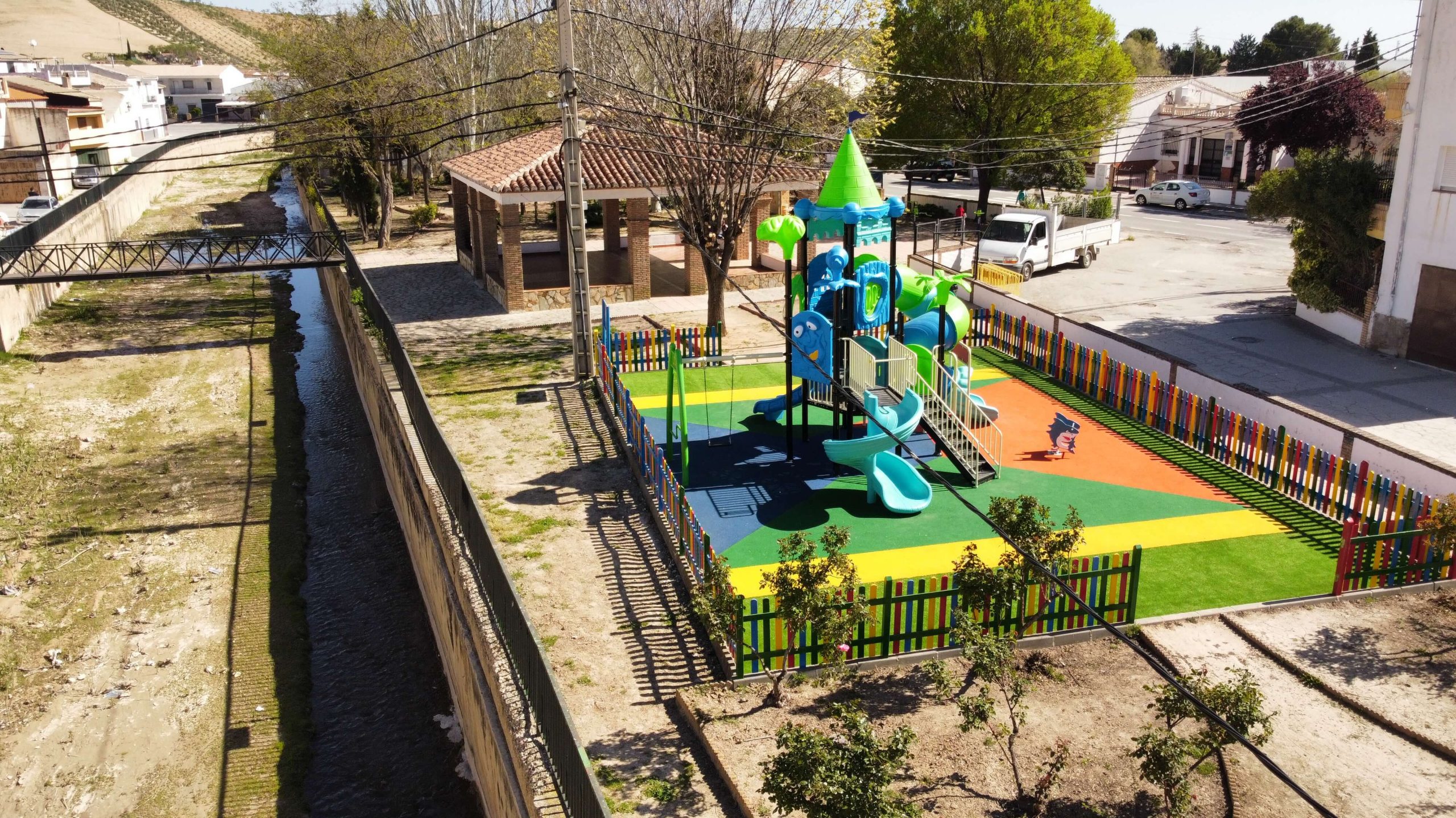 Parque infantil vallado con suelo de colores vibrantes y un gran conjunto de juegos con temática de castillo. La estructura principal es azul y verde, con toboganes, escaleras y plataformas de juego. Rodeado por una valla de madera pintada en colores llamativos, el parque está ubicado en una zona residencial con casas blancas y verdes, árboles y un puente en el fondo.