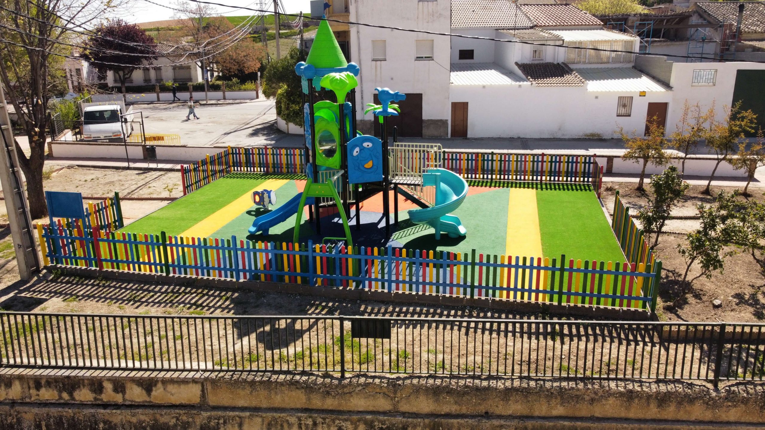Parque infantil vallado con suelo de colores vibrantes y un gran conjunto de juegos con temática de castillo. La estructura principal es azul y verde, con toboganes, escaleras y plataformas de juego. Rodeado por una valla de madera pintada en colores llamativos, el parque está ubicado en una zona residencial con casas blancas y verdes, árboles y un puente en el fondo.