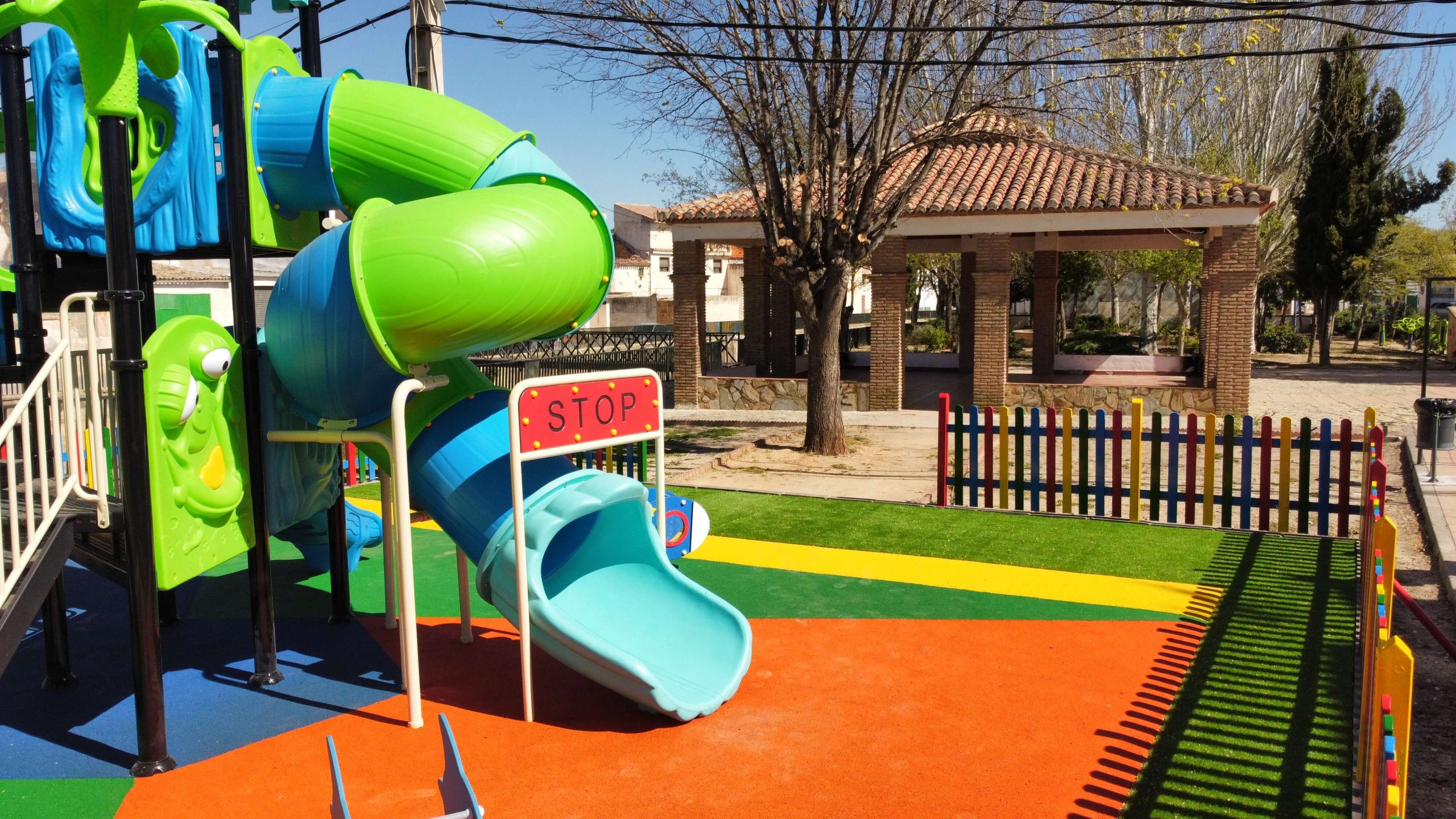 Parque infantil vallado con suelo de colores vibrantes y un gran conjunto de juegos con temática de castillo. La estructura principal es azul y verde, con toboganes, escaleras y plataformas de juego. Rodeado por una valla de madera pintada en colores llamativos, el parque está ubicado en una zona residencial con casas blancas y verdes, árboles y un puente en el fondo.
