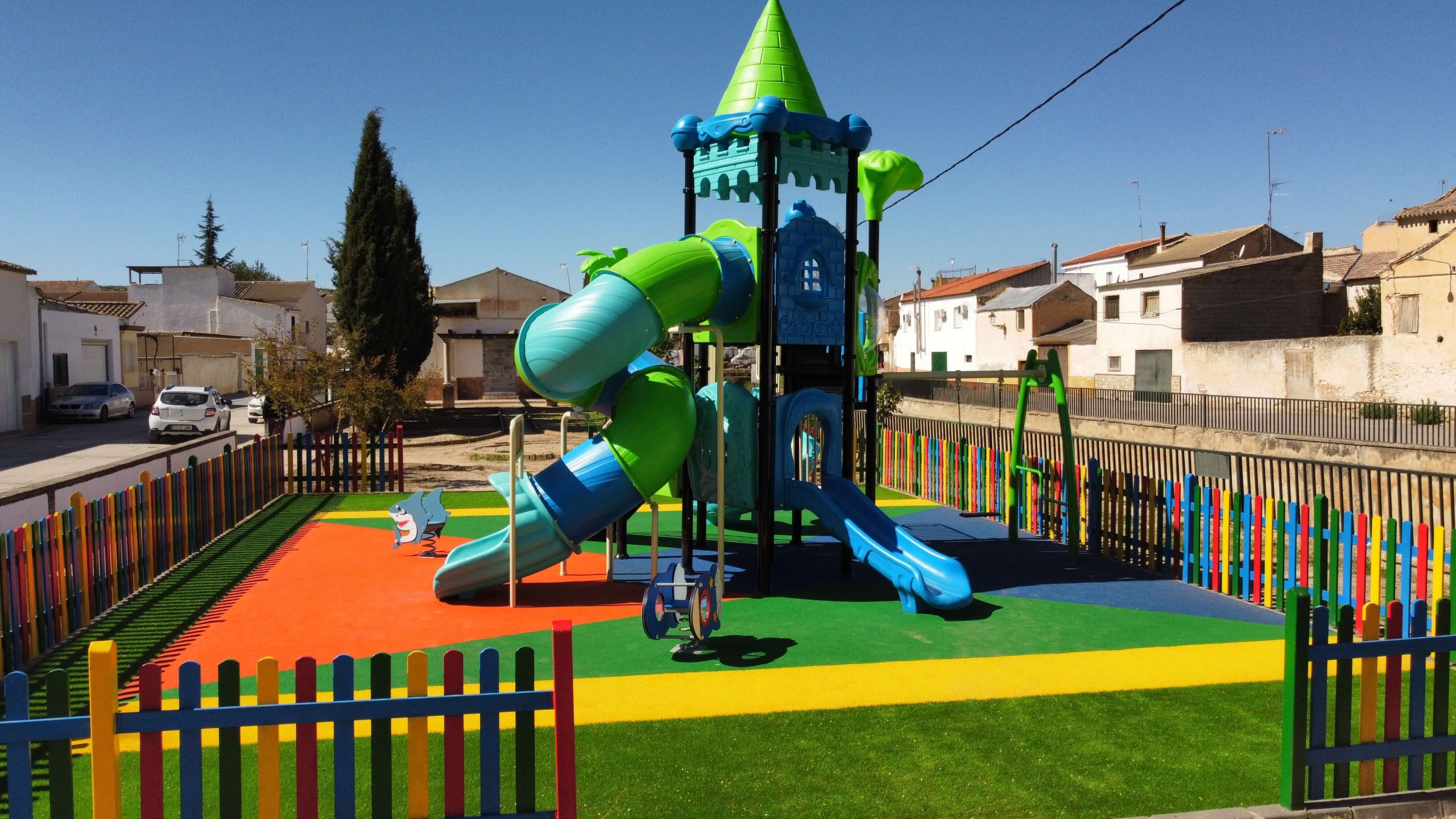 Parque infantil vallado con suelo de colores vibrantes y un gran conjunto de juegos con temática de castillo. La estructura principal es azul y verde, con toboganes, escaleras y plataformas de juego. Rodeado por una valla de madera pintada en colores llamativos, el parque está ubicado en una zona residencial con casas blancas y verdes, árboles y un puente en el fondo.