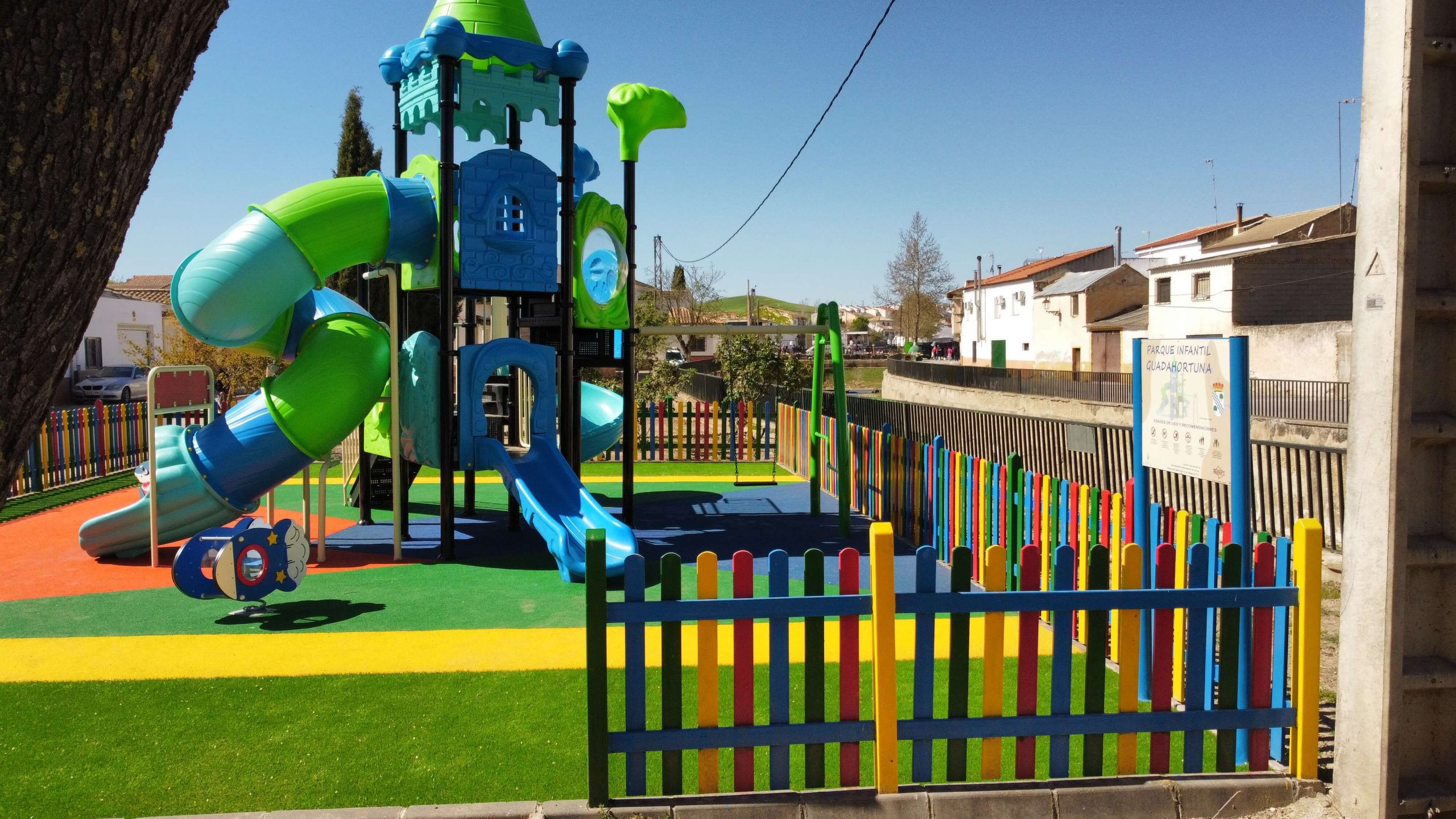 Parque infantil vallado con suelo de colores vibrantes y un gran conjunto de juegos con temática de castillo. La estructura principal es azul y verde, con toboganes, escaleras y plataformas de juego. Rodeado por una valla de madera pintada en colores llamativos, el parque está ubicado en una zona residencial con casas blancas y verdes, árboles y un puente en el fondo.