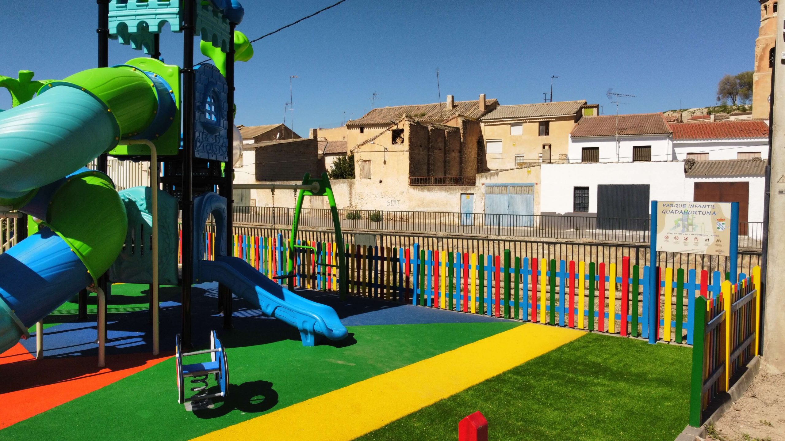 Parque infantil vallado con suelo de colores vibrantes y un gran conjunto de juegos con temática de castillo. La estructura principal es azul y verde, con toboganes, escaleras y plataformas de juego. Rodeado por una valla de madera pintada en colores llamativos, el parque está ubicado en una zona residencial con casas blancas y verdes, árboles y un puente en el fondo.