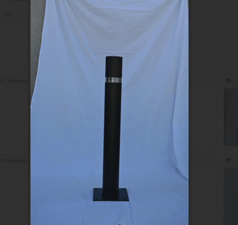 Pilona metálica negra con cuerpo cilíndrico liso, anillo superior de acero inoxidable y fijación atornillable al suelo.